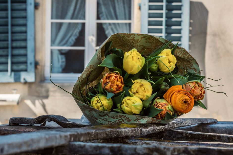 A bouquet of fresh flowers resting on a wrought iron surface outdoors, featuring bright yellow tulips, orange ranunculus, and orange and yellow peonies wrapped in decorative paper with green foliage i