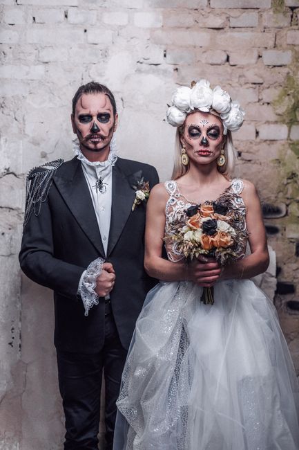 Two individuals dressed in elaborate costumes inspired by Día de los Muertos, standing against a textured brick wall. The person on the left wears a formal black tuxedo with lace details on the cuffs