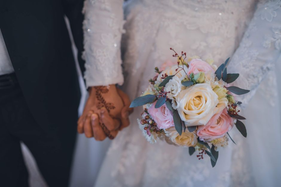 A close-up view of a couple holding hands during a wedding, with the bride holding a floral bouquet consisting of large white roses, soft pink roses, and small filler blooms, accented with eucalyptus 