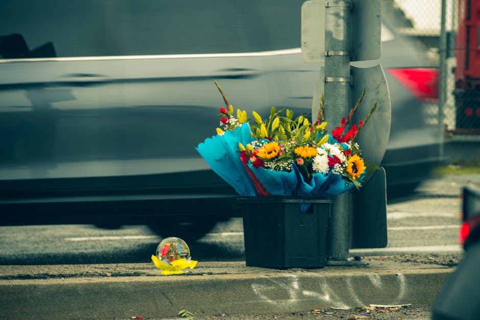 A black plastic delivery basket containing a colourful floral bouquet with yellow sunflowers, red and white flowers, and green foliage, wrapped in bright blue paper. The bouquet is placed next to a me