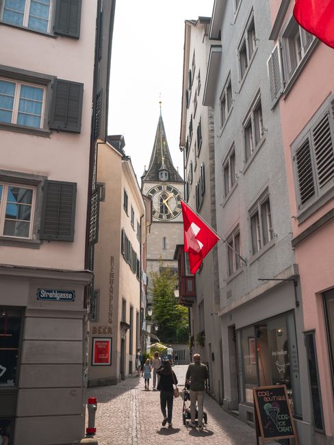 The image depicts a narrow European street view with tall, closely spaced buildings featuring light-colored facades and green shutters. In the background, a historic clock tower with a steep, pointed 