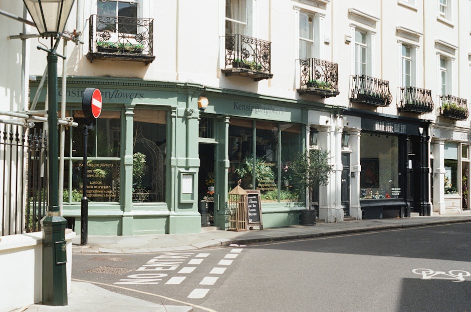 A picturesque street scene featuring a floral shop named Kensington Flowers housed in a classic Victorian building with a soft green facade, large display windows showcasing fresh flowers and plants, 