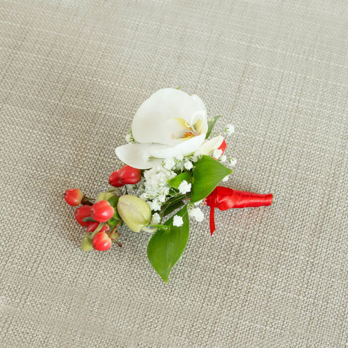 Set against a softly textured, light beige linen-style background that feels reminiscent of the calm, residential streets of De Beauvoir Town, this elegant boutonniere is photographed from above to highlight every considered detail. At its center lies a pristine white Phalaenopsis orchid, the petals broad, smooth, and gently curved, creating a graceful, wing-like form. In the heart of the orchid, a tender blend of creamy yellow and subtle orange adds depth and warmth, suggesting both purity and quiet celebration. Surrounding the orchid, fine sprays of baby's breath cluster in airy white tufts, each tiny blossom adding volume without heaviness, like a delicate veil of mist framing the main flower. Rich green foliage in varied shapes-some leaves sleek and pointed, others more rounded and compact-fans out from beneath the flowers, providing a natural structure and the sense of something freshly picked from a garden courtyard off De Beauvoir Road. Nestled along one side, a compact group of glossy, round red berries gleams brightly, the saturated hue offering a festive, almost jewel-like contrast to the white and green. Intermixed with them, a few pale green buds suggest new growth and seasonal freshness, ideal for winter weddings or holiday events. The stems of the boutonniere are neatly bound in a vivid red satin ribbon, tightly wrapped and secured with a small, secure knot whose short tails rest tidily on the fabric. Every element-the cool orchid, the feathery baby's breath, the polished berries, and the soft textile backdrop-contributes to a refined floral accessory designed to be worn close to the heart at a wedding, prom, or elegant celebration in and around De Beauvoir Town.